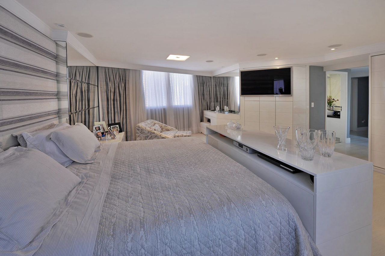 O amplo quarto do casal prima pelos tons claros. Na cabeceira os arquitetos optaram pela aplicação de papel de parede e espelhos. O teto rebaixado em gesso facilitou o uso de spots de iluminação e do sistema de som do home theater.