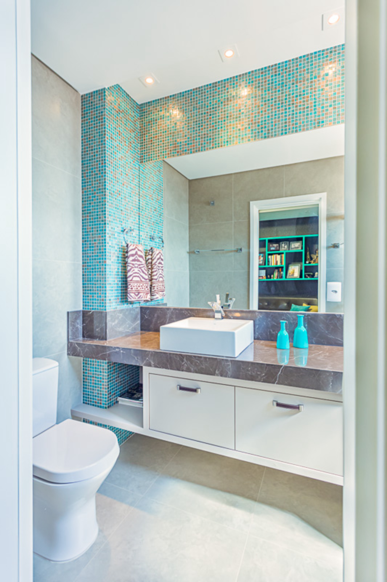 No banheiro que compõe o ambiente, pastilhas de vidro turquesa e bronze combinando com os nichos, para ficar em harmonia, e cimento queimado no piso e na  parede. 