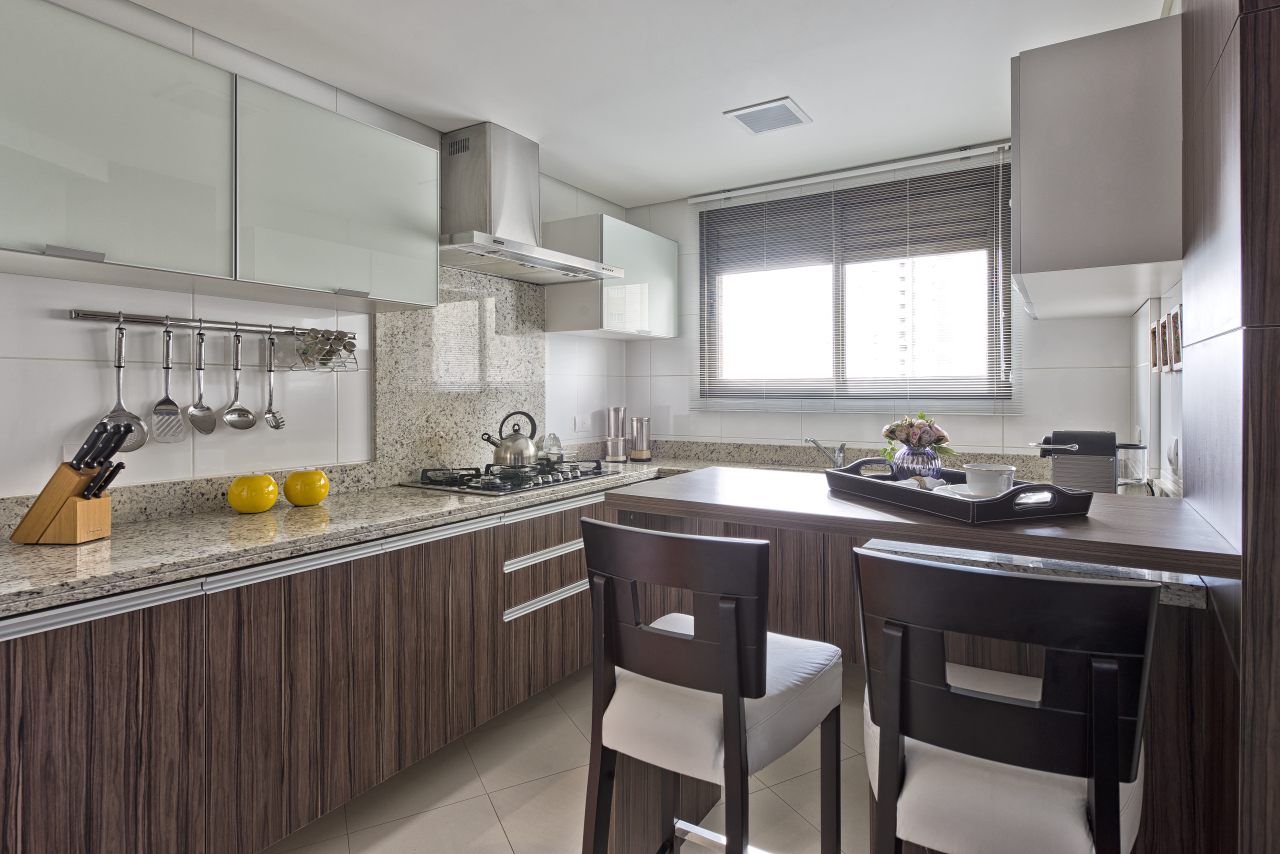 Na cozinha, em continuidade a sala de jantar, foi utilizado o tom madeirado escuro nos armários e branco criando contraste com os eletrodomésticos. Uma bancada central foi criada para descontrair na hora do preparo e facilitar o dia a dia.