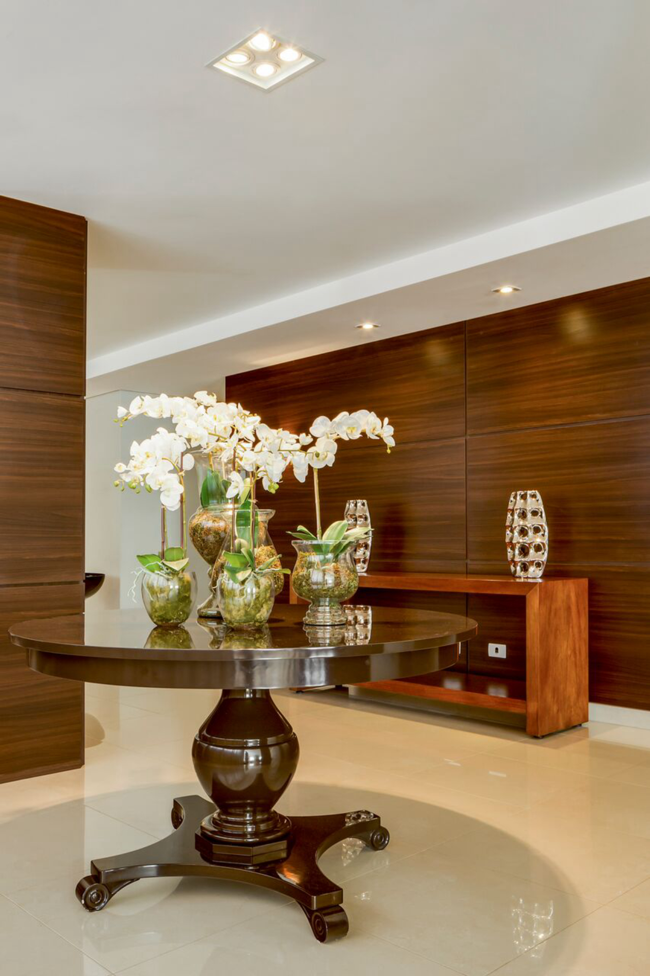 Os painéis de madeira oferecem conforto visual, ao mesmo tempo que aquecem o ambiente. A mesa central fica ainda mais em evidência com a iluminação direcionada. 