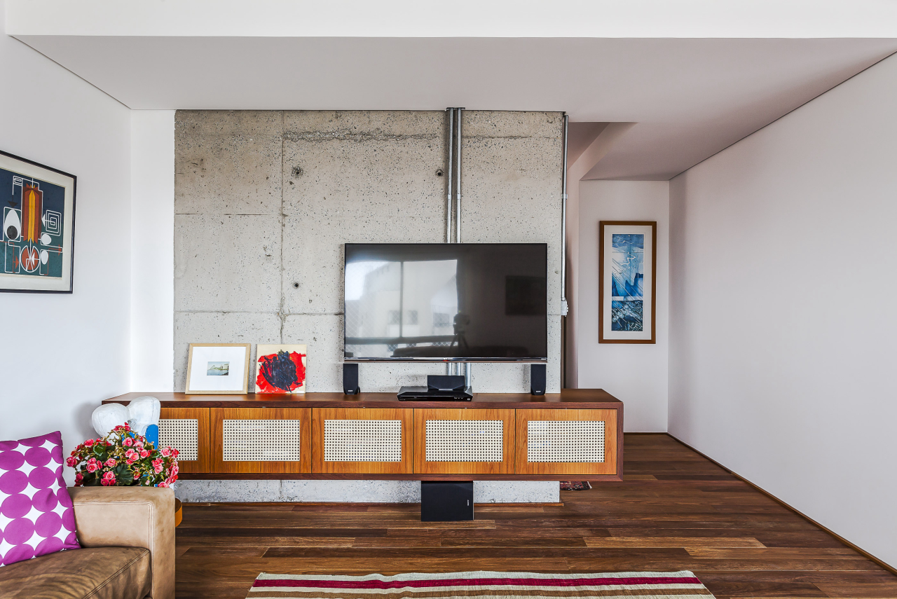 Área de tevê tem madeira  no piso e concreto na parede, conferindo ar moderno ao ambiente. 