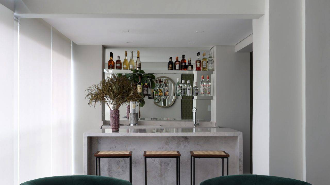 Um generoso bar foi montado com bancada de mármore e prateleiras espelhadas, em um lounge despojado com poltrona e mesas de apoio. 