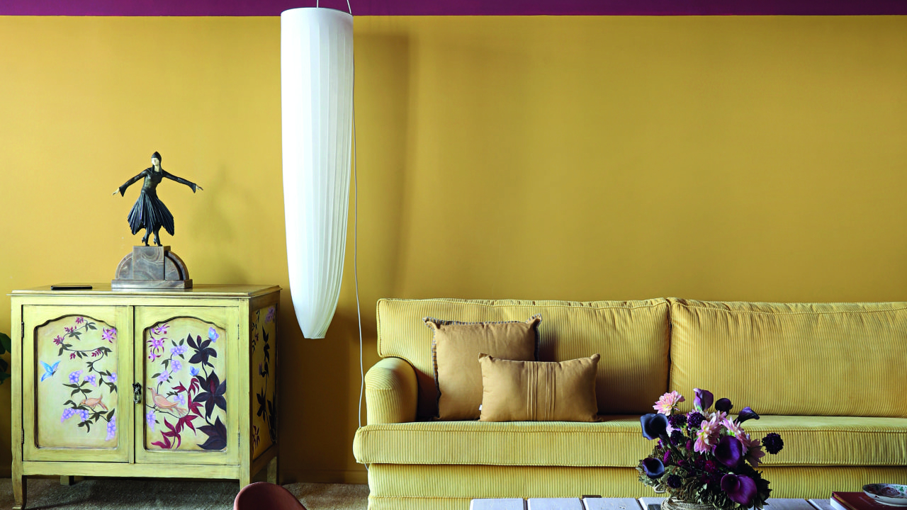 No living do apartamento de Lucila, a faixa amarela mimetiza o tom do sofá, que foi reformado com um novo tecido. A faixa superior da parede e o teto foram pintados de vermelho queimado para harmonizar as cores e integrar os dois ambientes. 